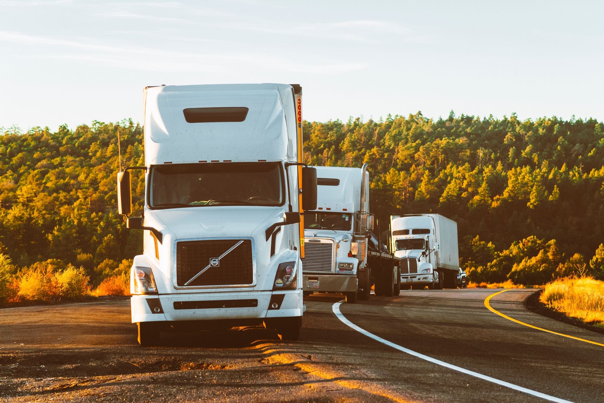 Three American trucks follow a curving highway, led by a white Volvo tractor with an enclosed box trailer visible behind the group.