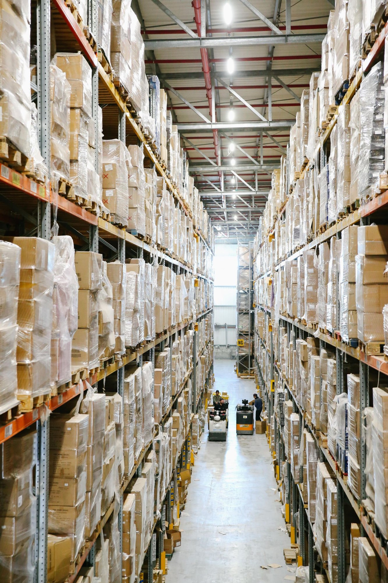Tall warehouse racks hold wrapped cartons beside an aisle with distant material-handling workers.