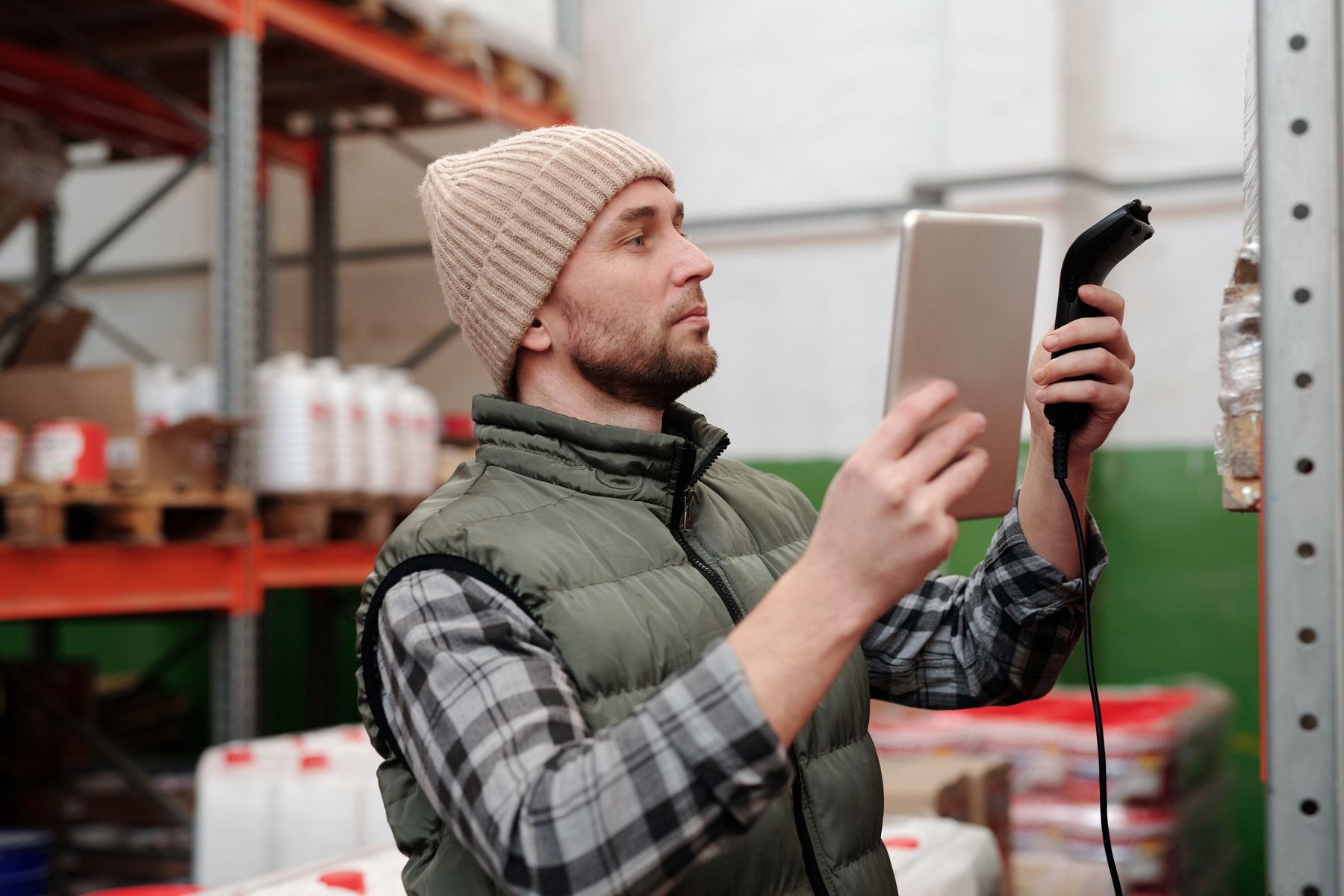 A warehouse worker in a knit cap checks inventory with a tablet and handheld scanner.